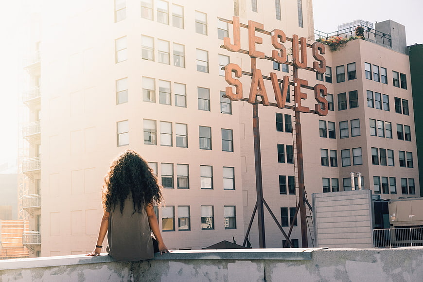 Woman with Curly Hair Sitting on Rooftop Facing Jesus Saves Sign