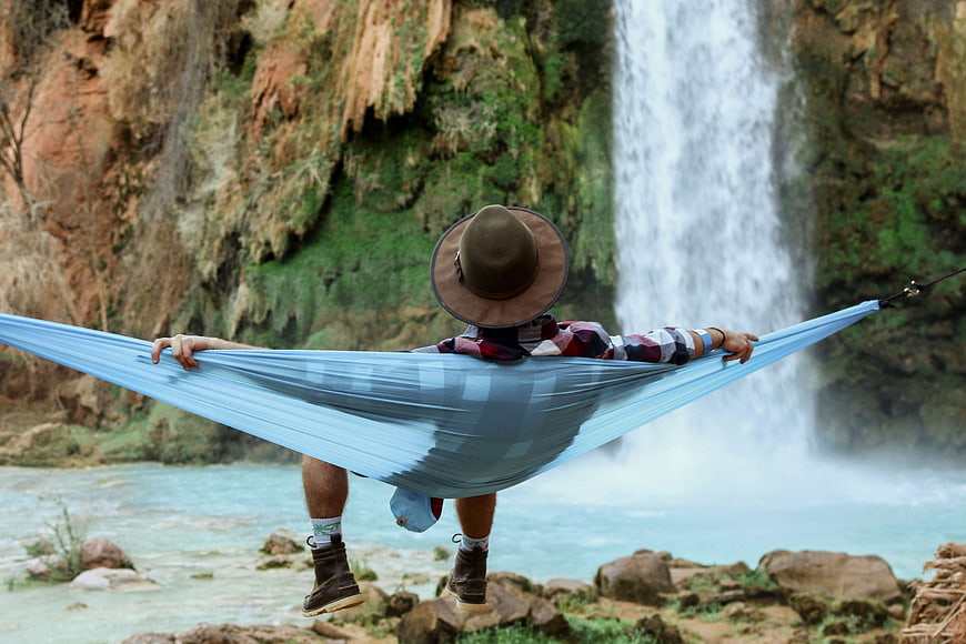 Man relaxing in blue hammock facing waterfall in nature