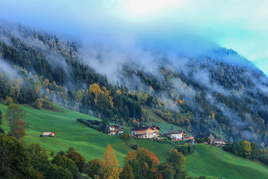 Misty Alpine Mountain Village With Green Pastures And Autumn Forest