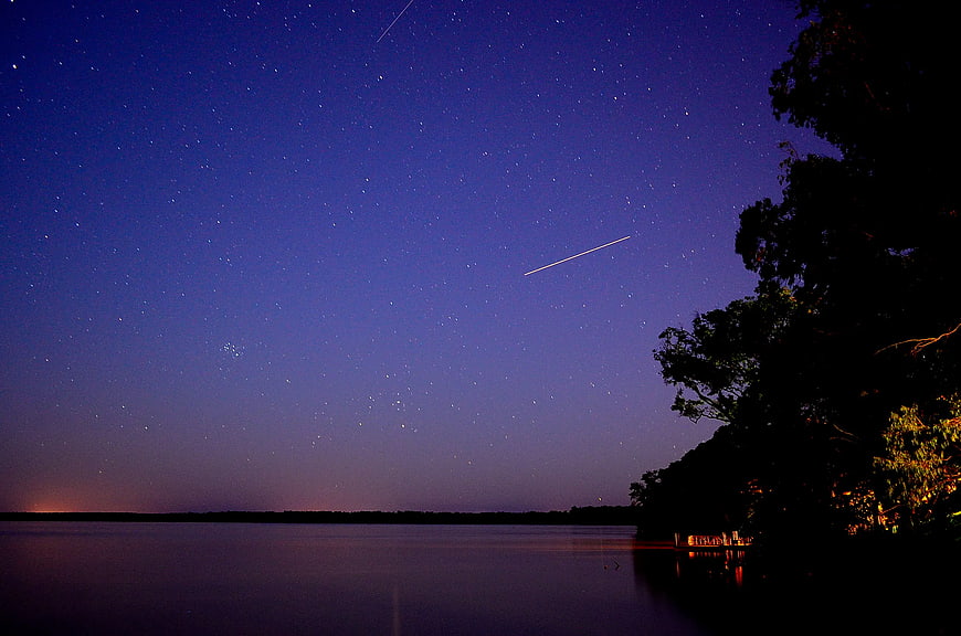 Meteor streaking across starry night sky over calm lake