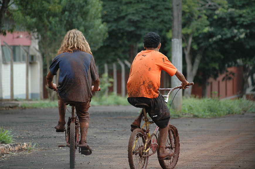 Two muddy children riding bicycles away on a dirt road