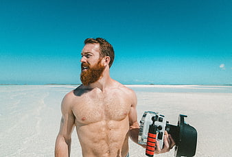 Shirtless bearded man holding camera on white sand beach