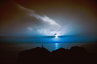 Silhouette of Person Standing on Rocky Cliff Overlooking Ocean at Dusk