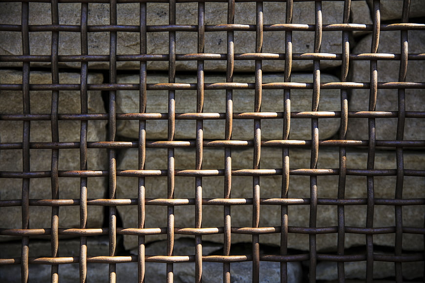 Close-up of rusted metal wire mesh fence against stone wall
