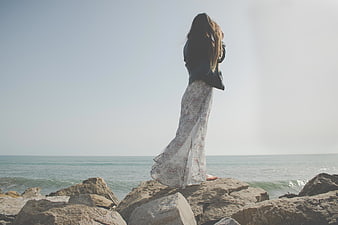 Woman in Long Dress Standing on Rocks by the Ocean
