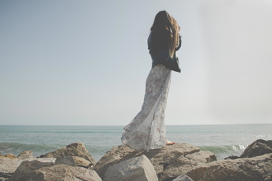 Woman in Long Dress Standing on Rocks by the Ocean