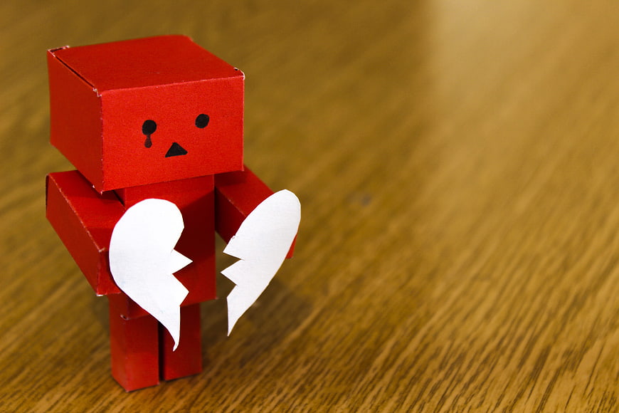 Sad red paper robot holding broken heart on wood