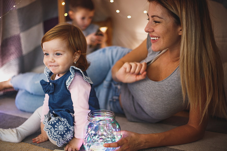 Happy mother and toddler playing with fairy lights in a cozy indoor tent