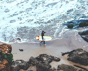 Aerial view of surfer walking on rocky beach with surfboard