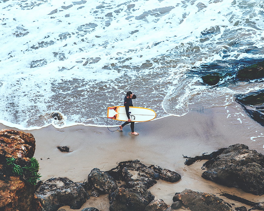 Aerial view of surfer walking on rocky beach with surfboard