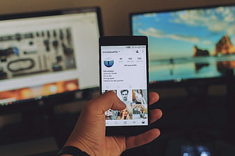 Hand holding smartphone with social media profile in front of computer monitors