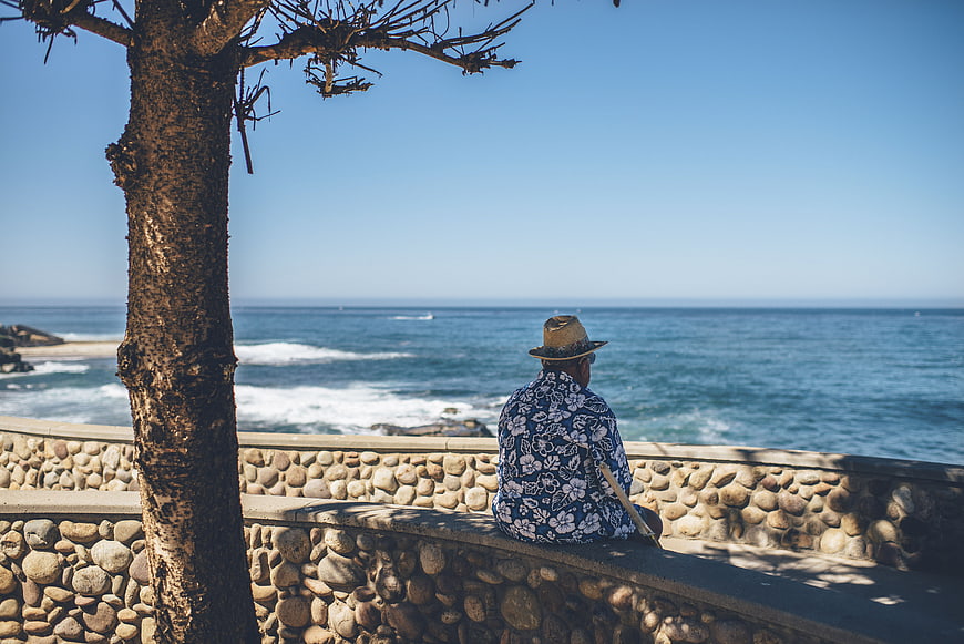 Man in Hawaiian Shirt Sitting on Stone Wall by Ocean