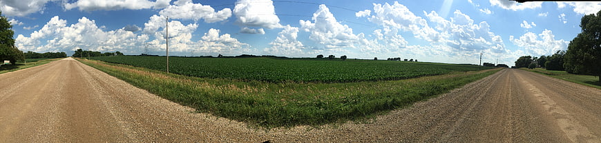 Panoramic rural dirt road intersection with green fields and blue sky
