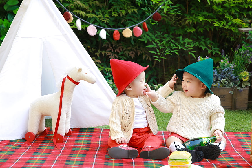 Two toddlers in gnome hats playing on a red plaid picnic blanket outdoors