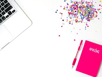 Flat lay of laptop, pink notebook, and colorful confetti on white background
