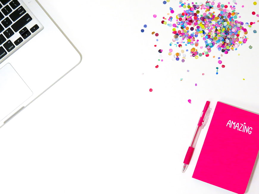 Flat lay of laptop, pink notebook, and colorful confetti on white background