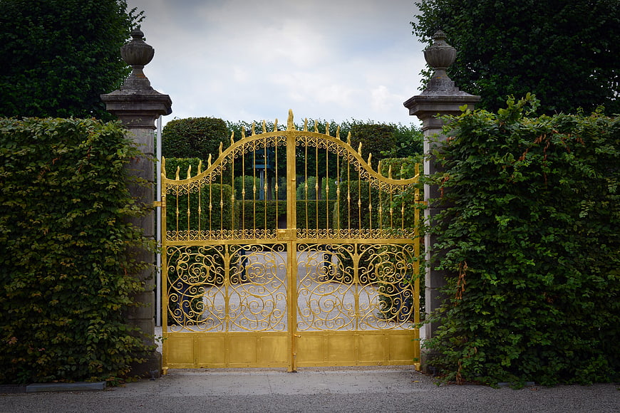Golden Ornate Wrought Iron Gate With Stone Pillars And Green Hedges