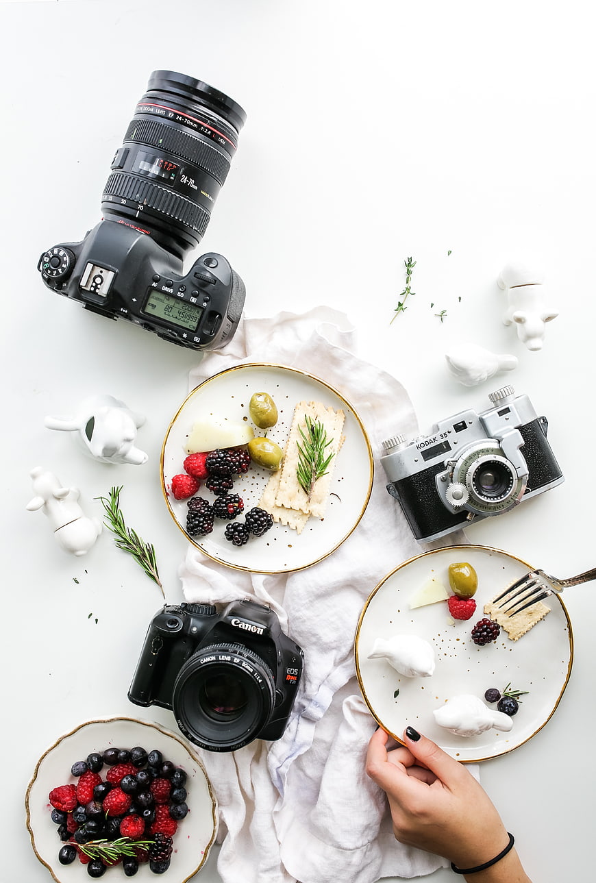 Flat lay of cameras, cheese, berries, and crackers on white background