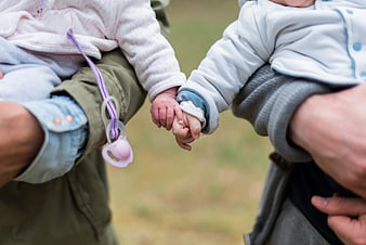 Two babies holding hands while being carried by parents outdoors