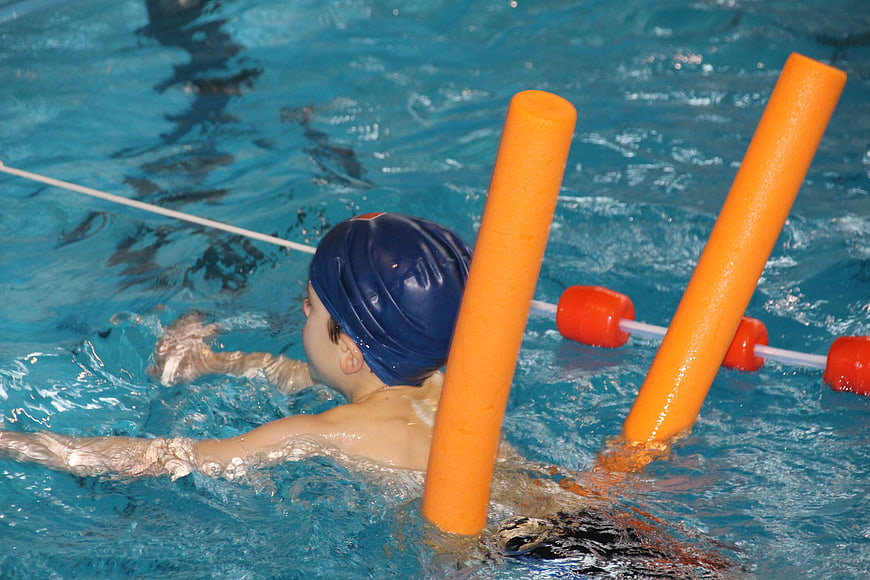 Young child swimming with orange pool noodle in blue water