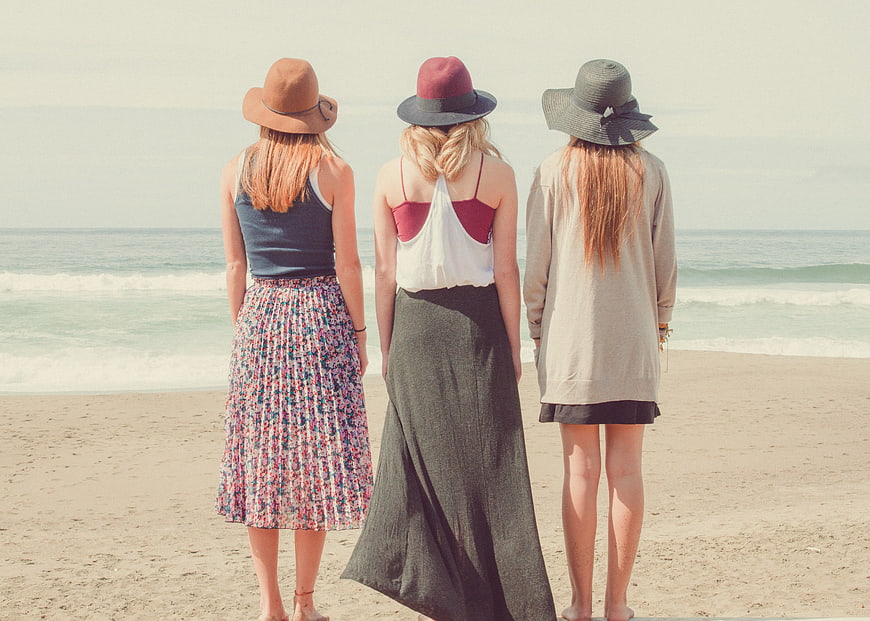 Three women in hats standing on sandy beach looking at ocean