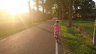 Young girl in pink dress riding bicycle on tree-lined road at golden hour