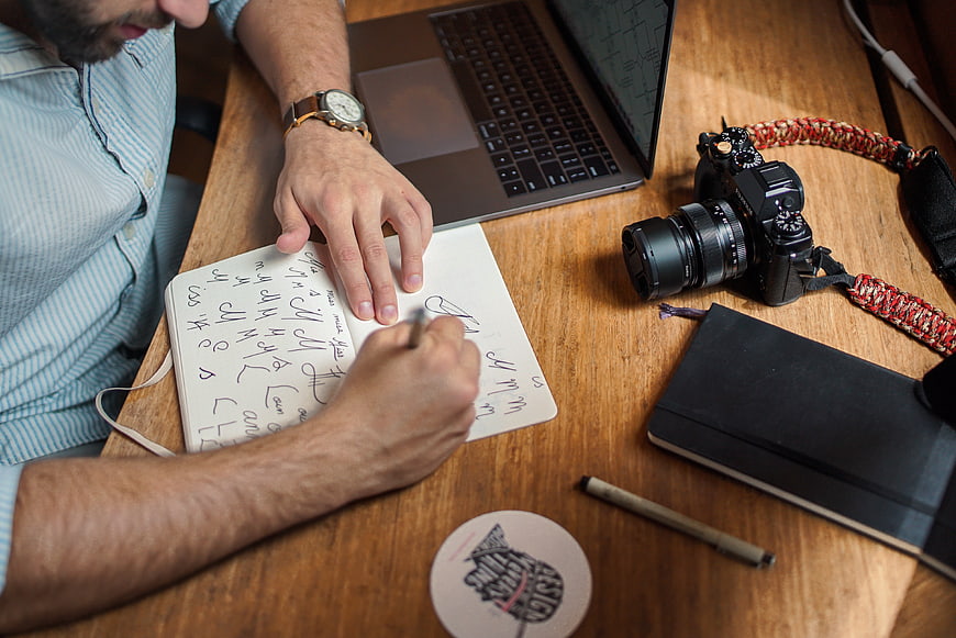 Man sketching design notes on desk with camera and laptop