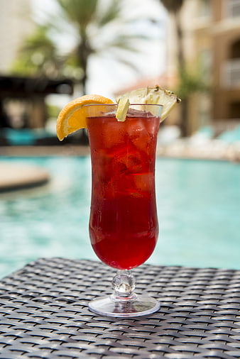 Red Cocktail With Orange And Pineapple Garnish On Wicker Table By Pool