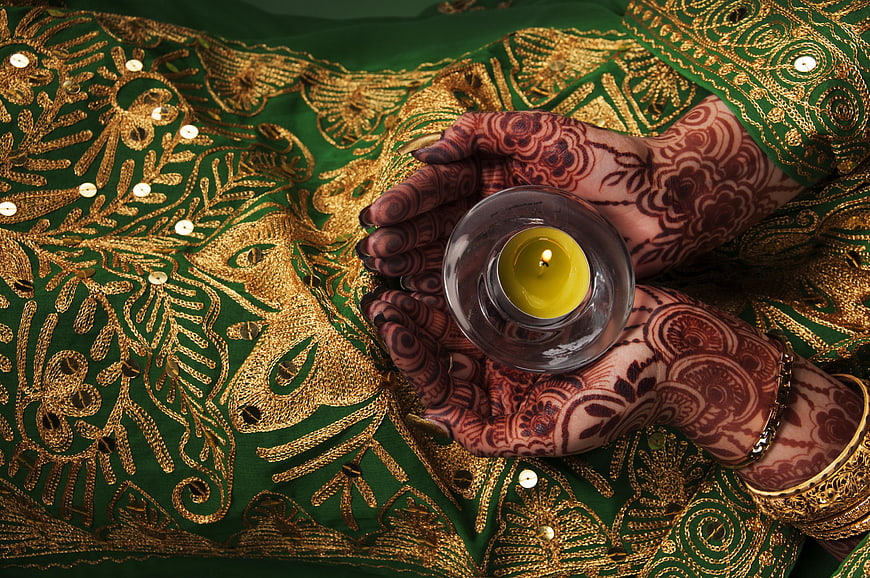 Henna-decorated hands holding a candle on green embroidered fabric