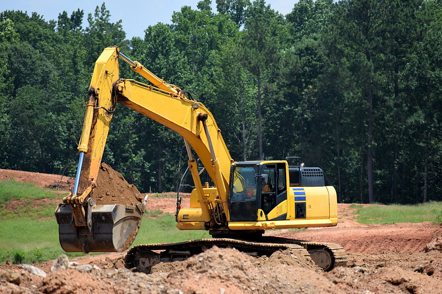 Yellow Excavator Digging Red Clay Soil In Forested Construction Site