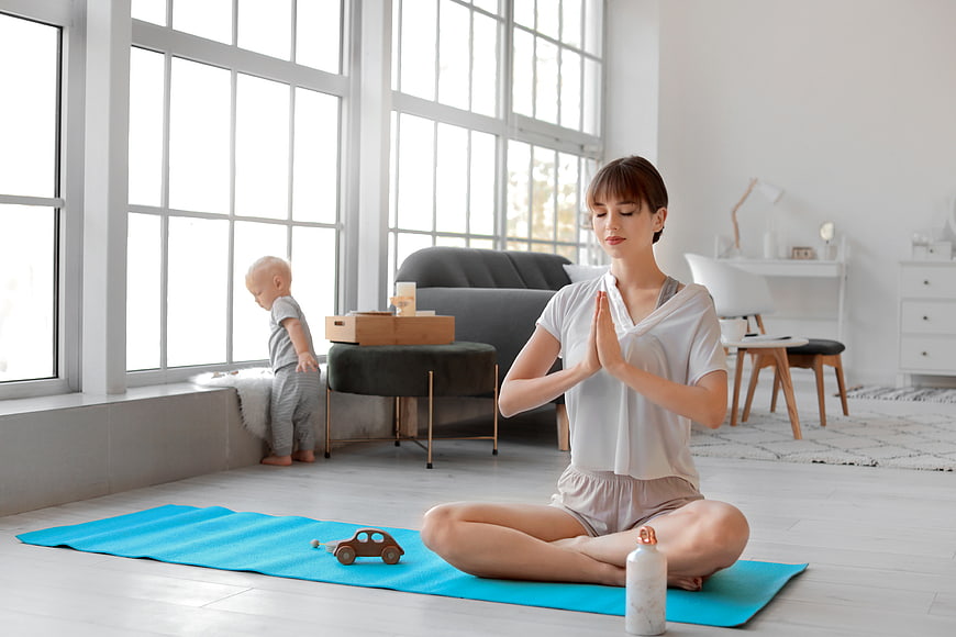Mother meditating at home with toddler playing nearby