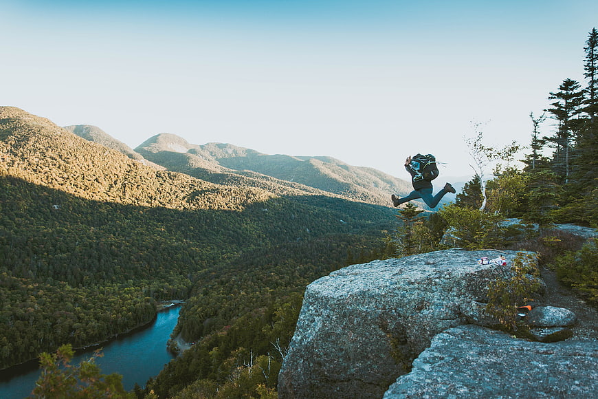 Hiker jumping off cliff with backpack in mountain landscape