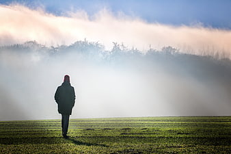 Lone figure walking in a foggy green field at dawn