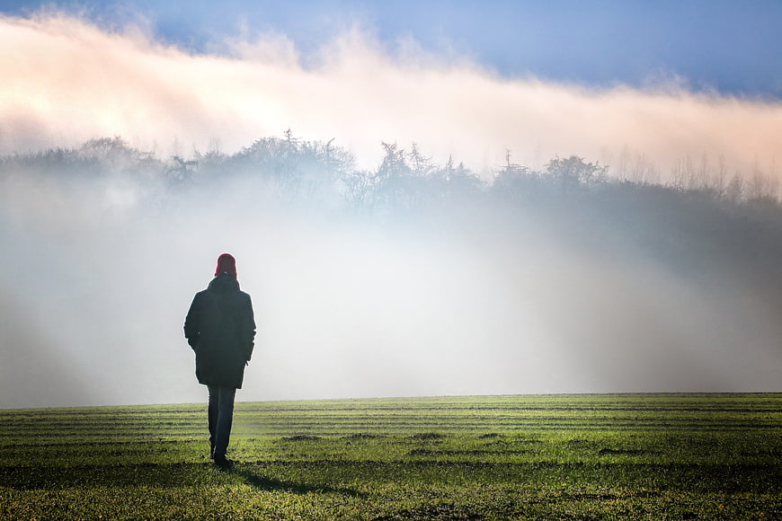 Lone figure walking in a foggy green field at dawn