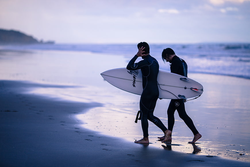 Two surfers walking on wet beach at dusk