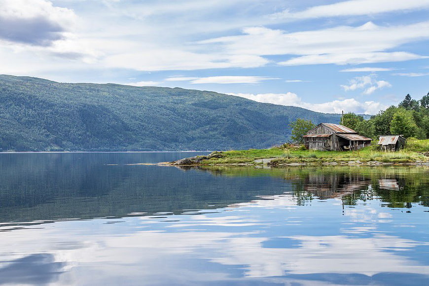Rustic wooden shack on calm lake shore with mountain backdrop