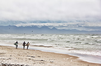 Surfers walking on sandy beach with mountains and ocean waves