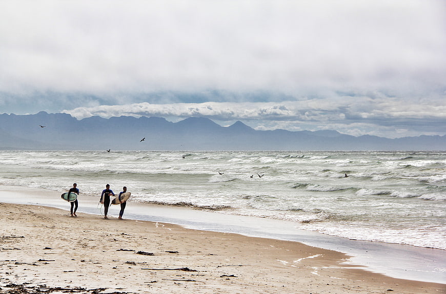 Surfers walking on sandy beach with mountains and ocean waves