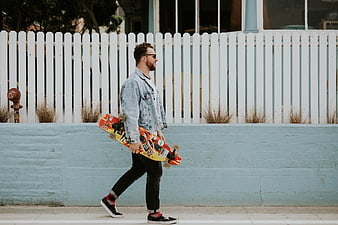 Man walking with skateboard along blue wall and white fence
