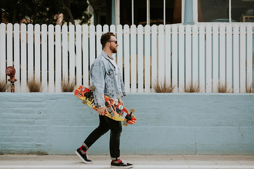 Man walking with skateboard along blue wall and white fence