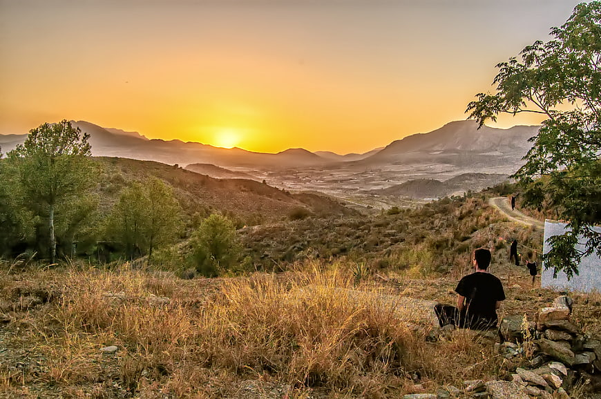 Man Sitting on Hillside Watching Golden Sunset Over Mountains