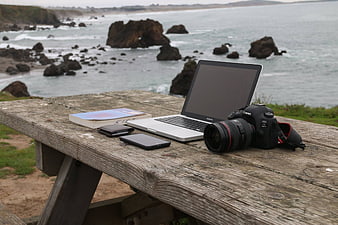 Photography gear and laptop on wooden table overlooking rocky ocean coast