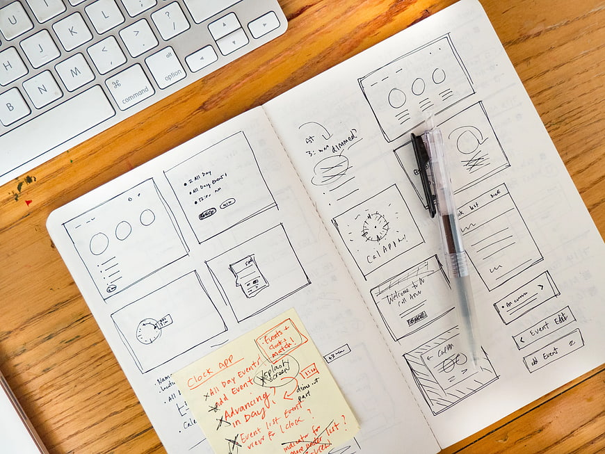Hand-drawn UI wireframes in sketchbook on wooden desk