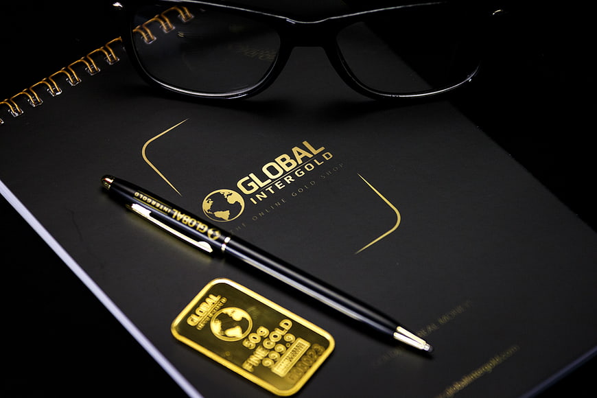 Black notebook with gold logo, pen, glasses, and gold bar