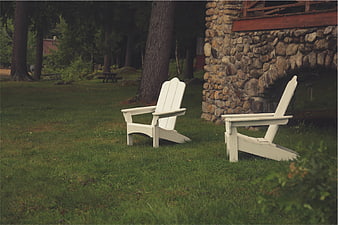 Two white Adirondack chairs on a grassy lawn near stone house