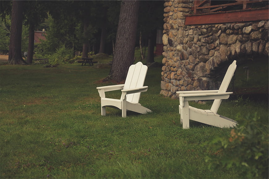 Two white Adirondack chairs on a grassy lawn near stone house