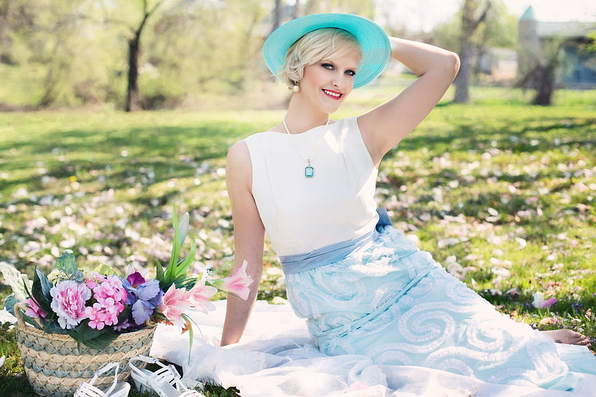 Blonde woman in blue hat and white dress sitting in spring park