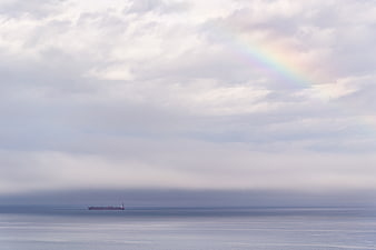 Cargo ship on calm ocean with faint rainbow in cloudy sky