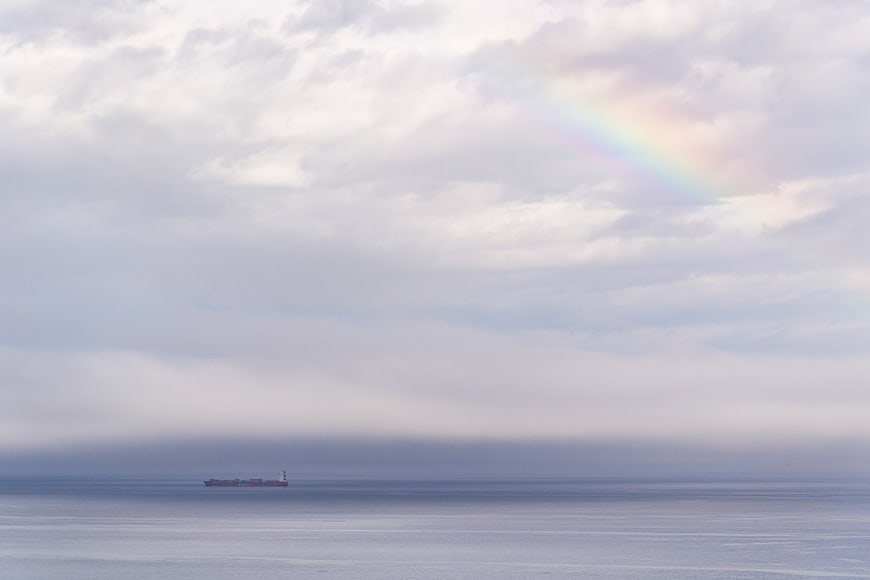 Cargo ship on calm ocean with faint rainbow in cloudy sky