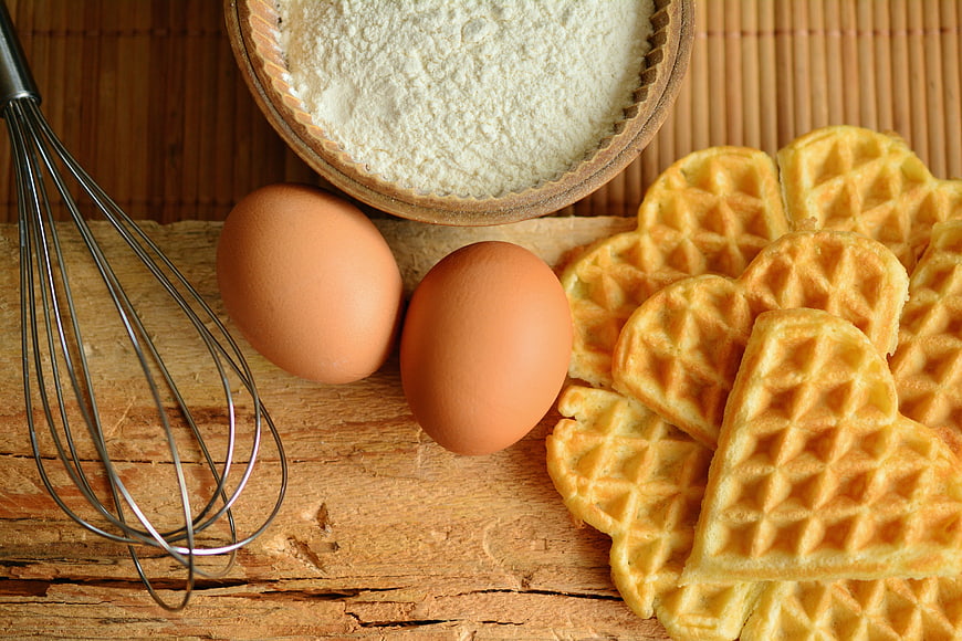Heart-shaped waffles with eggs, flour, and whisk on rustic wood